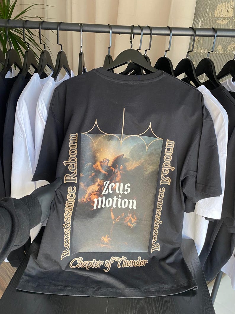 May include: Black t-shirt with a Renaissance-style graphic featuring the text "Zeus Motion" and "Chapter of Thunder." The design includes an image of a classical painting with figures. The shirt is hanging on a rack with other black and white shirts.