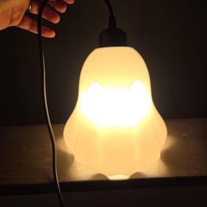 May include: A glowing, white ghost-shaped lamp with two eye-shaped cutouts. The lamp is lit from within, casting a warm light. A black cord extends from the top, held by a hand. The lamp sits on a dark surface.