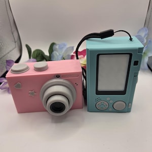 May include: A pink digital camera with a gray lens, two top buttons, and small heart and star accents, positioned next to a light blue digital device featuring a rectangular screen, a directional pad, and a circular button with a black wrist strap.