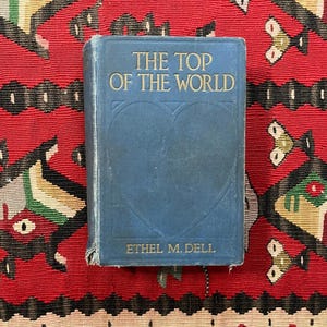 May include: A vintage book titled "The Top of the World" by Ethel M. Dell. The book has a blue cover with gold lettering and a heart-shaped design. The book is set against a red patterned background.