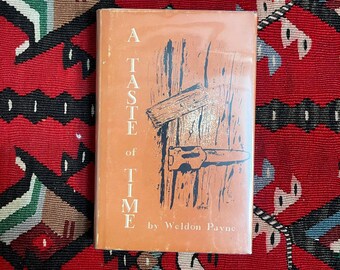 Puede incluir: Un libro antiguo titulado "A Taste of Time" de Weldon Payne. El libro tiene una portada naranja con una ilustración con temática de madera y el título en blanco. El libro está sobre una alfombra estampada roja y negra.