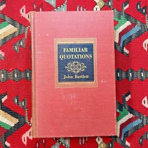 May include: A vintage book titled "Familiar Quotations" by John Bartlett. The book has a red cover with a blue and gold rectangular design in the center. The book is on a red patterned surface.