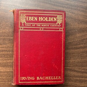Puede incluir: Un libro rojo vintage titulado "Eben Holden: A Tale of the North Country" de Irving Bacheller. La portada presenta letras doradas y elementos decorativos, con algo de desgaste visible en los bordes.