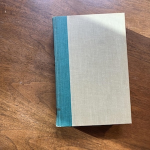 Puede incluir: Un libro de tapa dura con una cubierta gris claro y un lomo turquesa. El libro es rectangular y parece estar cerrado. El libro está sobre una superficie de madera.