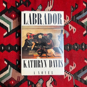 Könnte beinhalten: Ein Buch mit dem Titel "LABRADOR" von Kathryn Davis, Untertitel "A Novel". Das Cover zeigt ein Gemälde eines Raumes mit Figuren. Das Buch liegt auf einem roten gemusterten Teppich.