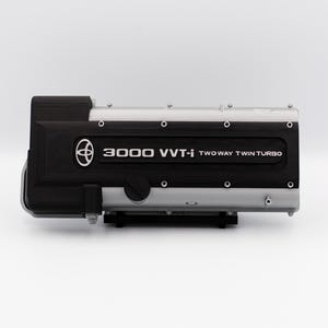 May include: A black and silver engine component with the text "3000 VVT-i TWO WAY TWIN TURBO" printed on it. The component has a Toyota logo and several circular fasteners. The engine part is displayed against a white background.