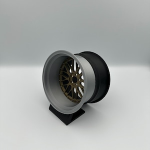 May include: A three-piece car wheel with a bronze-colored center, a gray rim, and a black outer section. The wheel is displayed on a black stand against a white background. The wheel's design suggests a focus on automotive customization.