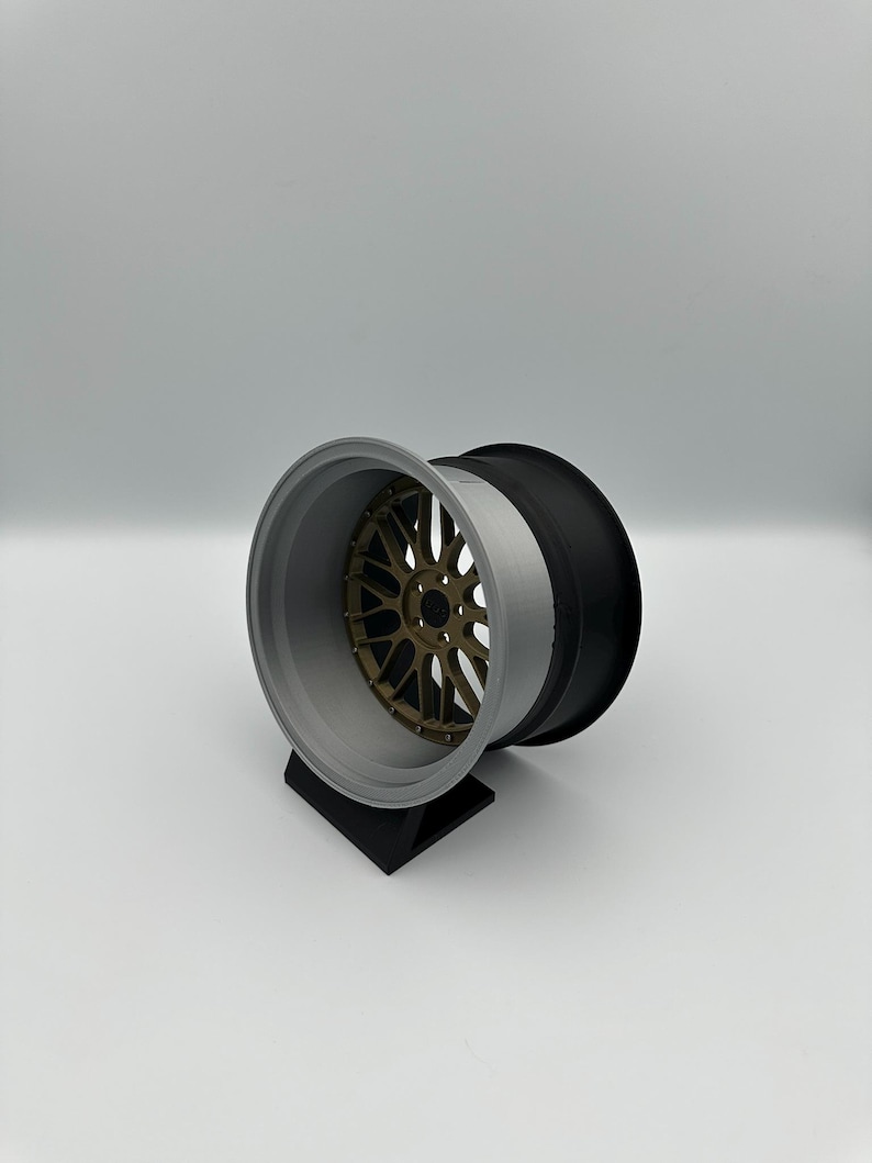 May include: A three-piece car wheel with a bronze-colored center, a gray rim, and a black outer section. The wheel is displayed on a black stand against a white background. The wheel's design suggests a focus on automotive customization.