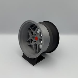以下が含まれることがあります： 黒とグレーの車のホイール。赤いセンターキャップが付いています。黒いスタンドに展示されています。ホイールはマルチスポークデザインとシルバーリムです。ホイールは3Dプリントモデルです。
