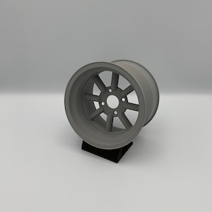 May include: A grey car wheel with eight spokes, displayed on a black stand. The wheel has a classic design, suitable for vintage or custom car builds. The background is plain white.