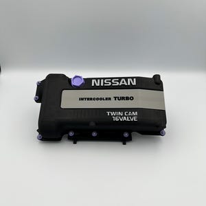 May include: Black engine cover with the word "NISSAN" in white. The cover has the words "INTERCOOLER TURBO" and "TWIN CAM 16VALVE" in white. The cover has purple accents.