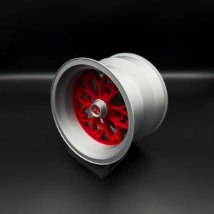 May include: A silver-colored wheel with a red, intricate, star-like design in the center. The wheel is set against a black background, showcasing its metallic finish and detailed design. The wheel is likely for a vehicle.