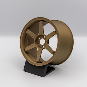 May include: A bronze-colored alloy wheel with a six-spoke design, displayed on a black stand. The wheel has a circular rim and a central hub with multiple bolt holes. The spokes have a textured surface, and the wheel is set against a white background.