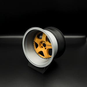May include: A car wheel with a gold-colored center and a silver rim. The tire is black. The wheel is displayed on a black stand against a black background. The center of the wheel has the text "LW0".