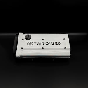Puede incluir: Una cubierta de motor plateada con el texto "TWIN CAM 20" y el logotipo de Toyota. La cubierta tiene un borde negro y varios pequeños sujetadores oscuros. El fondo es negro.