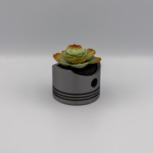 May include: A grey metal piston repurposed as a planter, holding a succulent with green and orange leaves. The cylindrical planter has a hole and horizontal grooves, creating a unique industrial-chic aesthetic. A creative and decorative item.
