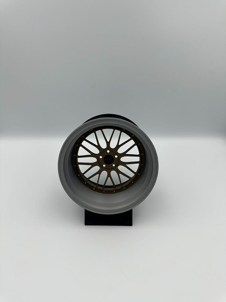 May include: A gray and bronze alloy wheel with a multi-spoke design. The wheel is displayed on a black stand against a white background. The wheel's rim is gray, and the spokes are bronze.