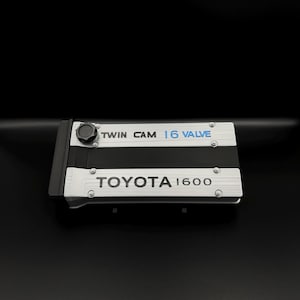 Puede incluir: Una cubierta de motor plateada y negra con el texto "TWIN CAM 16 VALVE" en blanco y azul, y "TOYOTA 1600" en blanco. La cubierta tiene una perilla negra en la parte superior.