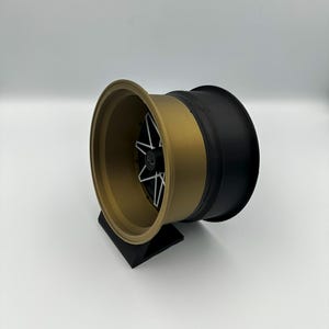 May include: A gold and black car wheel rim with a black center and a five-spoke design. The wheel is displayed on a black stand against a white background. The rim has a gold outer edge and a black inner edge.