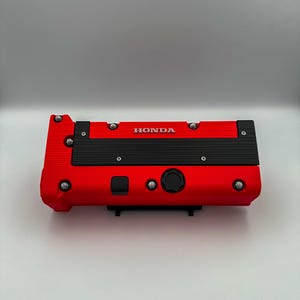 Puede incluir: Una tapa de válvula de motor roja con la palabra "HONDA" en letras blancas. La tapa tiene detalles en negro y varios pernos plateados. Probablemente sea una pieza de automóvil.