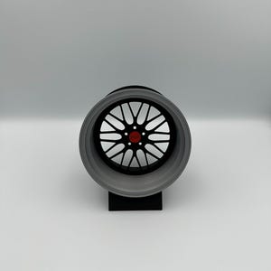 May include: A detailed, close-up view of a car wheel. The wheel features a black center with a complex spoke design and a red center cap. The outer rim is a light gray color. The wheel is mounted on a black stand against a white background.