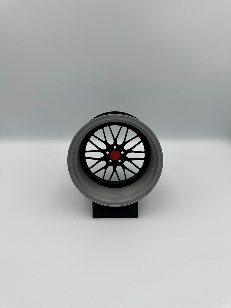 May include: A detailed, close-up view of a car wheel. The wheel features a black center with a complex spoke design and a red center cap. The outer rim is a light gray color. The wheel is mounted on a black stand against a white background.