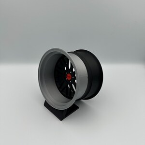 May include: A two-tone car wheel with a black center and a gray outer rim. The wheel is mounted on a black stand. The center of the wheel has a red accent.