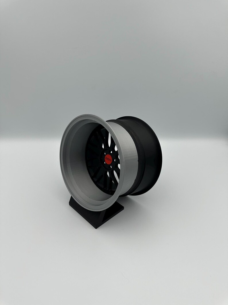 May include: A two-tone car wheel with a black center and a gray outer rim. The wheel is mounted on a black stand. The center of the wheel has a red accent.