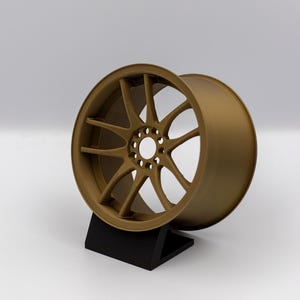 May include: A bronze alloy wheel with a multi-spoke design, presented on a black stand against a white backdrop. The wheel's structure includes numerous curved spokes radiating from the central hub to the outer rim, showcasing a detailed design.