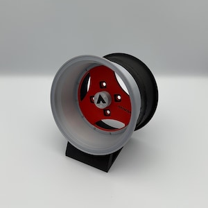 May include: A silver and red car wheel with a black center. The wheel has a three-spoke design with the word "ADVAN" visible. The wheel is displayed on a black stand against a white background.