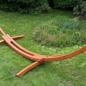 Puede incluir: Un soporte de hamaca de madera marrón con un diseño curvo. El soporte tiene una cadena en cada extremo para colgar una hamaca. Está colocado sobre un césped.
