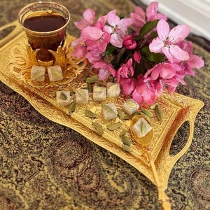 May include: A gold-colored, ornate serving tray holds a glass of tea, sweets, and pink flowers. The tray has a detailed design and handles. The tea is in a decorative glass. The sweets are square-shaped with green accents.