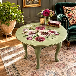 May include: A round sage green wooden coffee table with cabriole legs, featuring a large, detailed floral decal on the tabletop. The design displays clusters of white, pink, and deep magenta peonies and dahlias with green leaves on a muted surface.