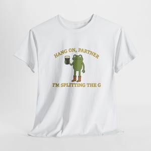 May include: White t-shirt with a cartoon frog holding a dark drink. The frog is wearing brown boots. The shirt has the text "HANG ON, PARTNER" above the frog and "I'M SPLITTING THE G" below. The text and frog are in shades of green and brown.
