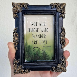 May include: A black framed art print with the quote "NOT ALL THOSE WHO WANDER ARE LOST" over a forest scene. The frame has ornate gold and black detailing. The artwork is held in a hand.