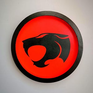 May include: Round, wooden wall art featuring the Thundercats logo. The design has a black border, a bright red center, and a black silhouette of the Thundercats emblem. The artwork is a vibrant and bold statement piece.