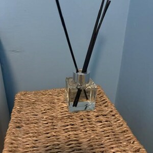 May include: A clear, square glass reed diffuser bottle with a silver-coloured collar. Several thin, black reed sticks are inserted into the bottle, extending upwards from the opening. The bottle contains a transparent liquid base for fragrance diffusion.