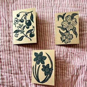 May include: Three blank greeting cards with black ink illustrations of flowers and berries. The cards are on a red and white striped fabric background.