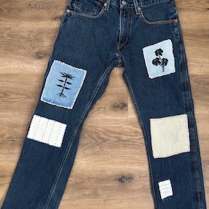 May include: Dark blue denim jeans with multiple patches. Patches include embroidered designs of a plant, a tree, and geometric patterns. The jeans have a classic five-pocket design and a button and zipper closure.