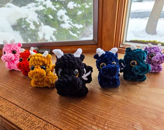 May include: A collection of seven small, plush, crocheted dragon figurines in various colours including pink, red, yellow, black, dark blue, teal, and purple. Each dragon features white horns, small wings, and large, glossy, dark-coloured eyes.