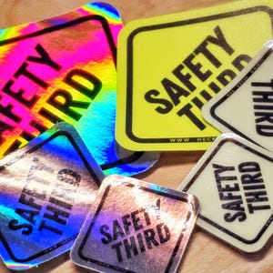 May include: A collection of square stickers with rounded corners, featuring the text "SAFETY THIRD." The stickers have various backgrounds, including iridescent, yellow, and off-white. The text is in black, and some stickers have the website address www.heck.fun.