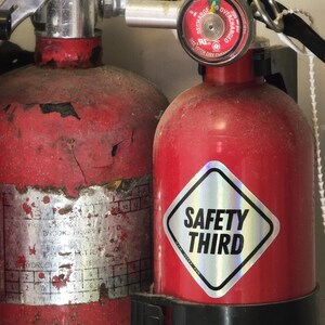 May include: Two red fire extinguishers. One is older with a worn label and the other has a silver and black diamond-shaped sticker that reads "SAFETY THIRD". The extinguishers are mounted in a black bracket.