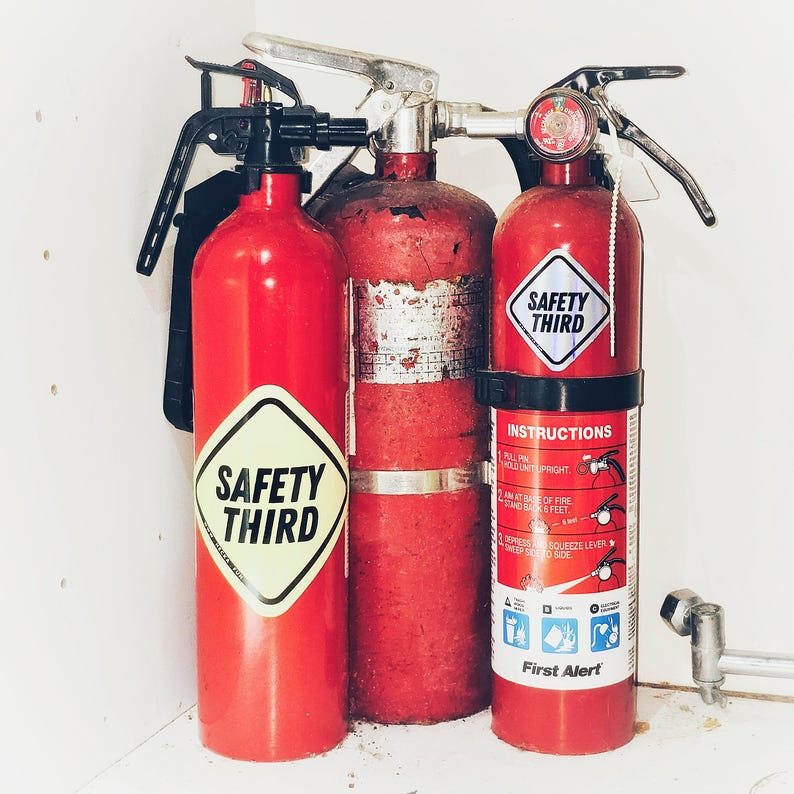 May include: A collection of red fire extinguishers with silver handles and black levers. Two extinguishers have "SAFETY THIRD" stickers, and one has a "First Alert" label with instructions. The extinguishers are mounted on a white surface.