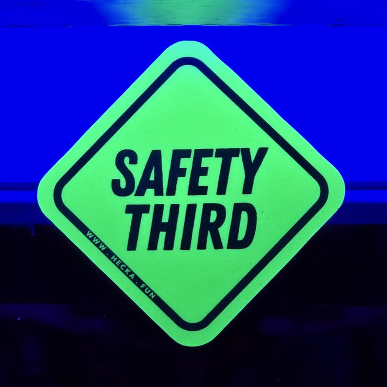 May include: A neon green diamond-shaped sticker with the words "SAFETY THIRD" in bold black letters. The sticker has a black border and the website address "WWW.HECKA.FUN" at the bottom. The background is a dark blue.