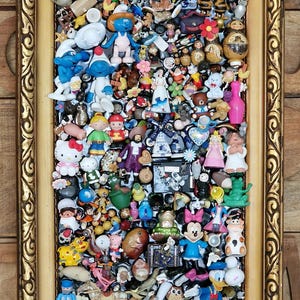 Könnte beinhalten: Ein goldfarbener Rahmen mit einer Sammlung verschiedener kleiner Figuren und Spielzeuge, darunter Zeichentrickfiguren, Puppen und Miniaturgegenstände. Der Rahmen hat eine verzierte Gestaltung, der Hintergrund ist eine Holztafel.