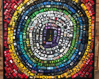 May include: A framed artwork featuring a spiral arrangement of toy cars in a rainbow colour pattern. The cars are arranged in concentric circles, with a traffic light in the centre. The frame is black.