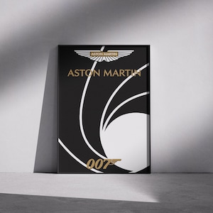 Puede incluir: Póster enmarcado en negro con el logotipo de Aston Martin en letras doradas y un diseño estilizado en blanco y negro. El texto "007" también es dorado. El póster está apoyado contra una pared gris.