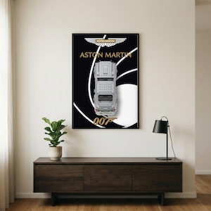 Puede incluir: Una lámina de arte mural enmarcada que muestra una vista cenital de un modelo de coche Aston Martin plateado sobre un fondo negro con gráficos circulares blancos, el logotipo de Aston Martin, el texto ASTON MARTIN y el texto dorado 007.
