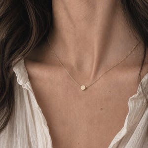 Könnte beinhalten: Eine zarte goldene Halskette mit einem kleinen, kreisförmigen Anhänger. Der Anhänger hängt an einer feinen Kette. Die Person trägt ein weißes Hemd mit Knöpfen. Ein schlichtes, elegantes Schmuckstück.