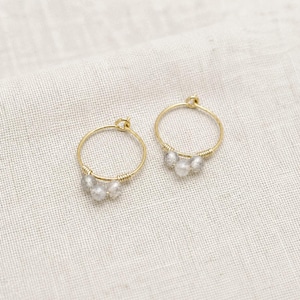 Peut inclure: Une paire de boucles d'oreilles créoles dorées. Chaque boucle d'oreille comporte un petit groupe de trois perles grises. Les perles sont fixées à la créole avec du fil doré. Les boucles d'oreilles sont présentées sur un tissu beige clair.